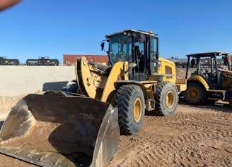 A large yellow construction vehicle, specifically a front loader, is parked on a dirt surface. The vehicle has large tires and a scooping shovel in front, which is slightly elevated. In the background, another similar construction vehicle and some industrial equipment or structures can be seen. The setting appears to be a construction site under a clear blue sky.