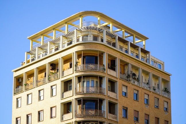 A multi-story building with yellow-beige brick exterior and rounded balcony edges. The upper floors feature large windows, multiple balconies adorned with potted plants, and a modern rooftop terrace with a pergola structure. The clear blue sky forms the backdrop.
