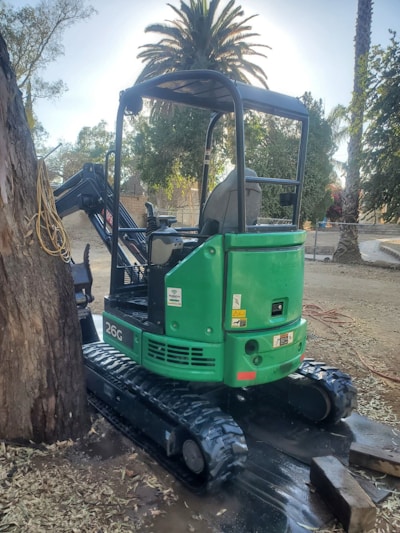 A green mini excavator with the model number 26G is parked next to a large tree. It features rubber tracks and an open operator's seat. The area surrounding the machine is outdoors with dirt ground, fallen leaves, and several tall palm trees in the background.