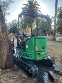 A green mini excavator with the model number 26G is parked next to a large tree. It features rubber tracks and an open operator's seat. The area surrounding the machine is outdoors with dirt ground, fallen leaves, and several tall palm trees in the background.