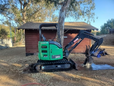 A green mini-excavator with a black arm and claw attachment is parked next to a large tree and a small wooden structure. The area is surrounded by trees and has a dirt ground with scattered sawdust or wood chips.
