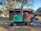 A green mini-excavator with a black arm and claw attachment is parked next to a large tree and a small wooden structure. The area is surrounded by trees and has a dirt ground with scattered sawdust or wood chips.
