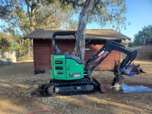 A green mini-excavator with a black arm and claw attachment is parked next to a large tree and a small wooden structure. The area is surrounded by trees and has a dirt ground with scattered sawdust or wood chips.