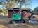 A green mini-excavator with a black arm and claw attachment is parked next to a large tree and a small wooden structure. The area is surrounded by trees and has a dirt ground with scattered sawdust or wood chips.