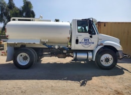 A white tanker truck featuring the logo 'UCL United Construction & Landscape' is parked on a dirt surface. The truck has large wheels and is positioned side-on, with shadows visible beneath it. In the background, there are trees and a large yellow container.