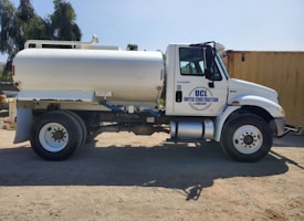 A white tanker truck featuring the logo 'UCL United Construction & Landscape' is parked on a dirt surface. The truck has large wheels and is positioned side-on, with shadows visible beneath it. In the background, there are trees and a large yellow container.