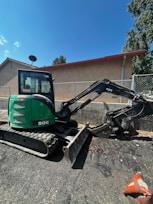 A green mini excavator with a large bucket is parked on a gravel surface near a chain-link fence. The machine has tracks instead of wheels and is positioned to one side. Behind it, there is a building with a light brown wall and a sloped red roof. A satellite dish is visible on the side of the building. Trees and the blue sky form the background.