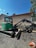 A green mini excavator with a large bucket is parked on a gravel surface near a chain-link fence. The machine has tracks instead of wheels and is positioned to one side. Behind it, there is a building with a light brown wall and a sloped red roof. A satellite dish is visible on the side of the building. Trees and the blue sky form the background.