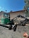 A green mini excavator with a large bucket is parked on a gravel surface near a chain-link fence. The machine has tracks instead of wheels and is positioned to one side. Behind it, there is a building with a light brown wall and a sloped red roof. A satellite dish is visible on the side of the building. Trees and the blue sky form the background.