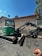 A green mini excavator with a large bucket is parked on a gravel surface near a chain-link fence. The machine has tracks instead of wheels and is positioned to one side. Behind it, there is a building with a light brown wall and a sloped red roof. A satellite dish is visible on the side of the building. Trees and the blue sky form the background.