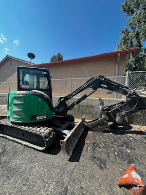 A green mini excavator with a large bucket is parked on a gravel surface near a chain-link fence. The machine has tracks instead of wheels and is positioned to one side. Behind it, there is a building with a light brown wall and a sloped red roof. A satellite dish is visible on the side of the building. Trees and the blue sky form the background.