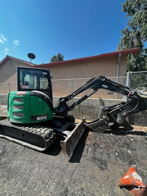 A green mini excavator with a large bucket is parked on a gravel surface near a chain-link fence. The machine has tracks instead of wheels and is positioned to one side. Behind it, there is a building with a light brown wall and a sloped red roof. A satellite dish is visible on the side of the building. Trees and the blue sky form the background.