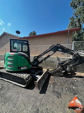 A green mini excavator with a large bucket is parked on a gravel surface near a chain-link fence. The machine has tracks instead of wheels and is positioned to one side. Behind it, there is a building with a light brown wall and a sloped red roof. A satellite dish is visible on the side of the building. Trees and the blue sky form the background.