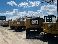 A line of large, yellow excavators featuring the CAT logo is parked on a gravel lot under a bright blue sky with scattered clouds. A billboard is visible in the background.