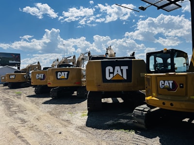 A line of large, yellow excavators featuring the CAT logo is parked on a gravel lot under a bright blue sky with scattered clouds. A billboard is visible in the background.