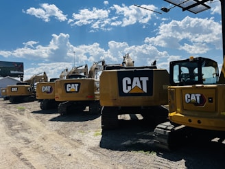 A line of large, yellow excavators featuring the CAT logo is parked on a gravel lot under a bright blue sky with scattered clouds. A billboard is visible in the background.
