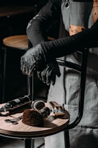 A barber or hair stylist stands with arms crossed, wearing a grey apron and black gloves. On a wooden stool in front are hairdressing tools including scissors, brushes, clippers, and a spray bottle.