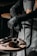 A barber or hair stylist stands with arms crossed, wearing a grey apron and black gloves. On a wooden stool in front are hairdressing tools including scissors, brushes, clippers, and a spray bottle.