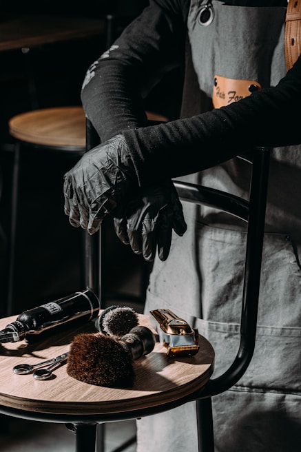 A barber or hair stylist stands with arms crossed, wearing a grey apron and black gloves. On a wooden stool in front are hairdressing tools including scissors, brushes, clippers, and a spray bottle.