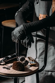 A barber or hair stylist stands with arms crossed, wearing a grey apron and black gloves. On a wooden stool in front are hairdressing tools including scissors, brushes, clippers, and a spray bottle.