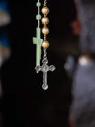 A close-up view of a rosary with a tiny metallic cross hanging alongside spherical golden beads. In the background, a blurred image of another cross is visible.