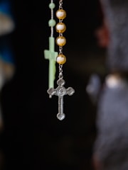 A close-up view of a rosary with a tiny metallic cross hanging alongside spherical golden beads. In the background, a blurred image of another cross is visible.