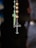 A close-up view of a rosary with a tiny metallic cross hanging alongside spherical golden beads. In the background, a blurred image of another cross is visible.
