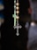 A close-up view of a rosary with a tiny metallic cross hanging alongside spherical golden beads. In the background, a blurred image of another cross is visible.