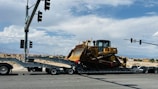A large yellow bulldozer is secured on a trailer, which is part of a multi-axle transport truck stopped at an intersection. Several traffic lights are visible above, while hills and a few buildings can be seen in the distant background under a partly cloudy sky.