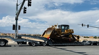 A large yellow bulldozer is secured on a trailer, which is part of a multi-axle transport truck stopped at an intersection. Several traffic lights are visible above, while hills and a few buildings can be seen in the distant background under a partly cloudy sky.
