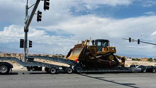 A large yellow bulldozer is secured on a trailer, which is part of a multi-axle transport truck stopped at an intersection. Several traffic lights are visible above, while hills and a few buildings can be seen in the distant background under a partly cloudy sky.