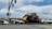 A large yellow bulldozer is secured on a trailer, which is part of a multi-axle transport truck stopped at an intersection. Several traffic lights are visible above, while hills and a few buildings can be seen in the distant background under a partly cloudy sky.