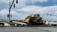 A large yellow bulldozer is secured on a trailer, which is part of a multi-axle transport truck stopped at an intersection. Several traffic lights are visible above, while hills and a few buildings can be seen in the distant background under a partly cloudy sky.