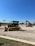 A construction equipment yard featuring several pieces of heavy machinery, including a large yellow bulldozer and an excavator under a clear blue sky. The ground is covered in gravel with a grassy edge in the foreground. In the background, there are more construction vehicles and a sign reading 'Holt CAT.'
