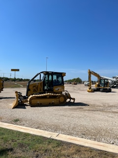 A construction equipment yard featuring several pieces of heavy machinery, including a large yellow bulldozer and an excavator under a clear blue sky. The ground is covered in gravel with a grassy edge in the foreground. In the background, there are more construction vehicles and a sign reading 'Holt CAT.'
