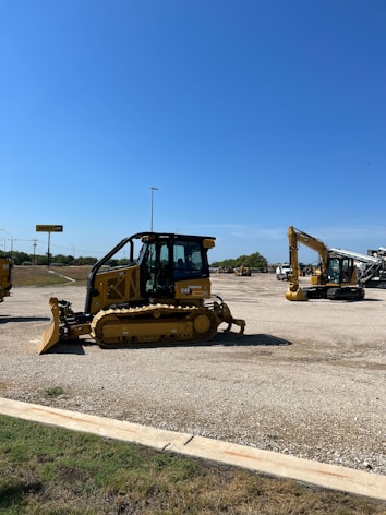 A construction equipment yard featuring several pieces of heavy machinery, including a large yellow bulldozer and an excavator under a clear blue sky. The ground is covered in gravel with a grassy edge in the foreground. In the background, there are more construction vehicles and a sign reading 'Holt CAT.'