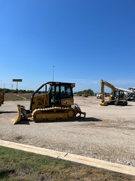 A construction equipment yard featuring several pieces of heavy machinery, including a large yellow bulldozer and an excavator under a clear blue sky. The ground is covered in gravel with a grassy edge in the foreground. In the background, there are more construction vehicles and a sign reading 'Holt CAT.'
