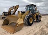 Two large construction vehicles are parked on a dirt surface under an overcast sky. The foreground features a yellow wheel loader with large treaded tires and a front bucket. In the background, there is an excavator with a visible boom and cab.