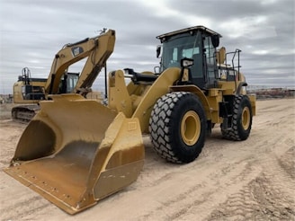 Two large construction vehicles are parked on a dirt surface under an overcast sky. The foreground features a yellow wheel loader with large treaded tires and a front bucket. In the background, there is an excavator with a visible boom and cab.
