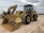 Two large construction vehicles are parked on a dirt surface under an overcast sky. The foreground features a yellow wheel loader with large treaded tires and a front bucket. In the background, there is an excavator with a visible boom and cab.