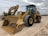 Two large construction vehicles are parked on a dirt surface under an overcast sky. The foreground features a yellow wheel loader with large treaded tires and a front bucket. In the background, there is an excavator with a visible boom and cab.