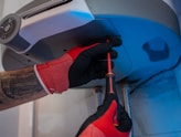 A person wearing red and black gloves is using a screwdriver to work on the underside of a metallic appliance. The appliance is mounted and has visible pipes connected to it. The person's forearm shows a tattoo.