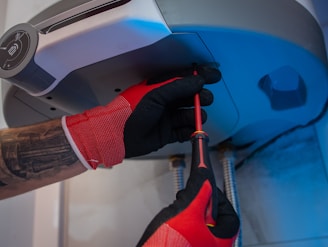 A person wearing red and black gloves is using a screwdriver to work on the underside of a metallic appliance. The appliance is mounted and has visible pipes connected to it. The person's forearm shows a tattoo.