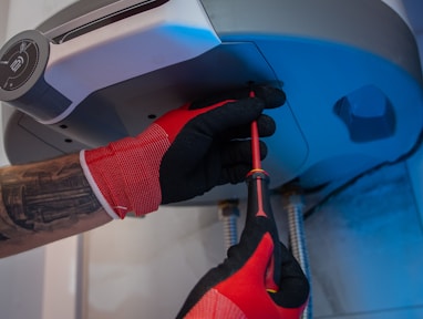 A person wearing red and black gloves is using a screwdriver to work on the underside of a metallic appliance. The appliance is mounted and has visible pipes connected to it. The person's forearm shows a tattoo.