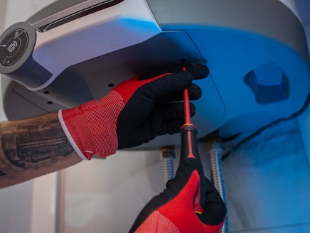 A person wearing red and black gloves is using a screwdriver to work on the underside of a metallic appliance. The appliance is mounted and has visible pipes connected to it. The person's forearm shows a tattoo.
