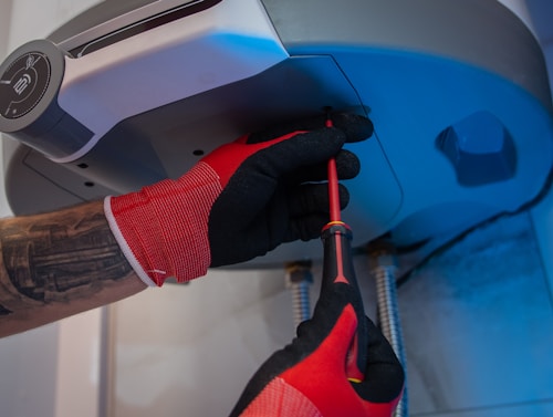 A person wearing red and black gloves is using a screwdriver to work on the underside of a metallic appliance. The appliance is mounted and has visible pipes connected to it. The person's forearm shows a tattoo.