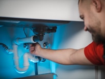 A person in a red shirt is fixing or inspecting plumbing under a sink. The scene includes various pipes, connectors, and a disposal unit. The lighting is bluish, illuminating the area where the person is working.