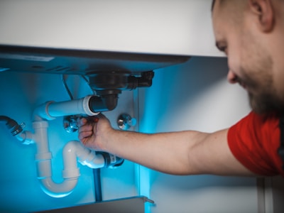 A person in a red shirt is fixing or inspecting plumbing under a sink. The scene includes various pipes, connectors, and a disposal unit. The lighting is bluish, illuminating the area where the person is working.
