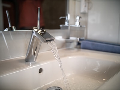 A modern bathroom sink with a sleek, metallic faucet flowing with clear water. The countertop is white, and there are accessories like soap dispensers visible in the background.