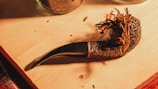 A wooden pipe with dried tobacco strands is placed on a wooden surface. The pipe has a textured, dark brown finish and a curved stem. Nearby, parts of glass jars are visible, suggesting a cozy setting.
